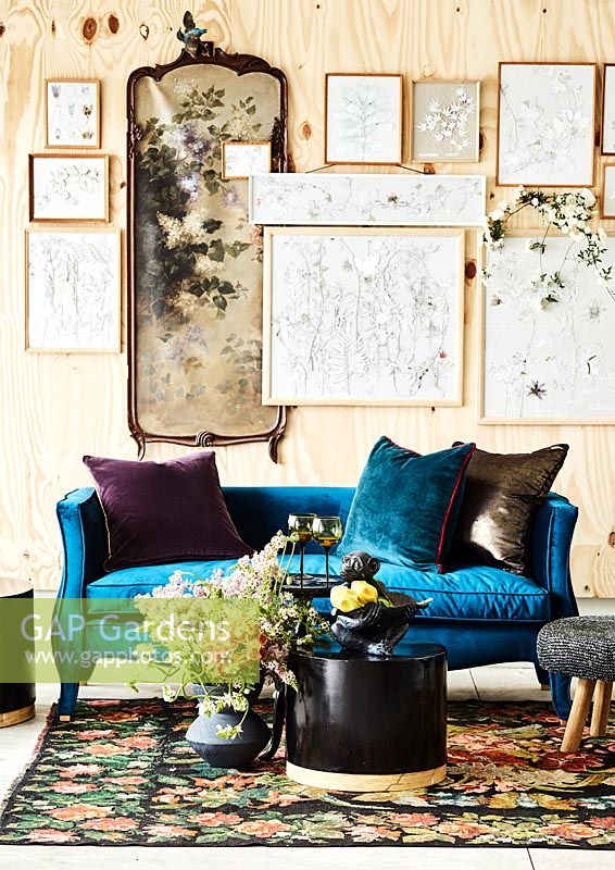 Un mur d'illustrations encadrées derrière un canapé bleu, une table et des fleurs fraîches.
