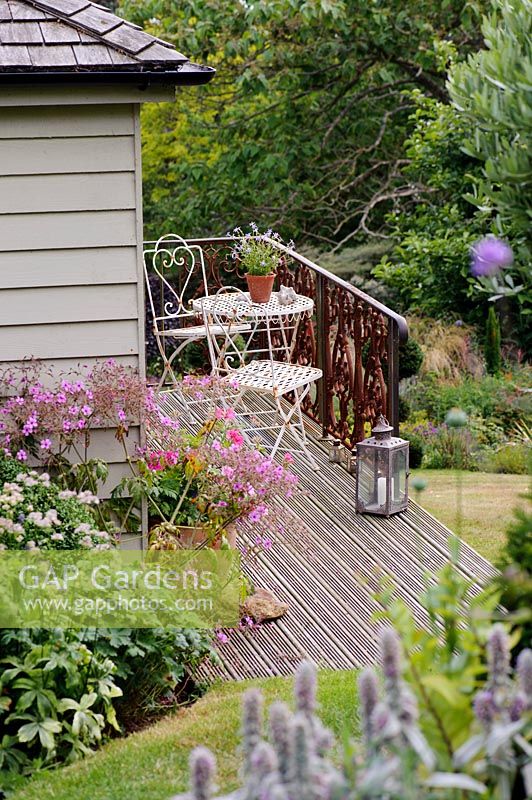 Vue de la table et des chaises de bistrot en métal blanc sur une terrasse en bois à l'extérieur du studio de jardin.