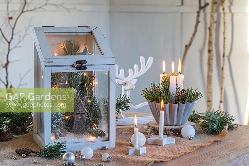 Arrangement festif utilisant du feuillage et des pommes de pin, des lumières LED, des bougies et des boules dans un cadre rustique.