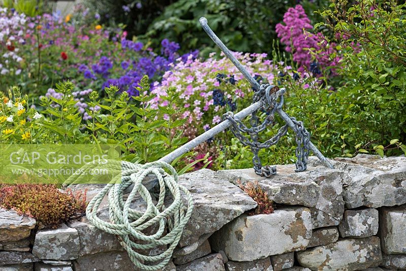 Une vieille ancre et perche de corde au sommet d'un mur en pierre naturelle à l'extérieur d'un jardin coloré en bord de mer.