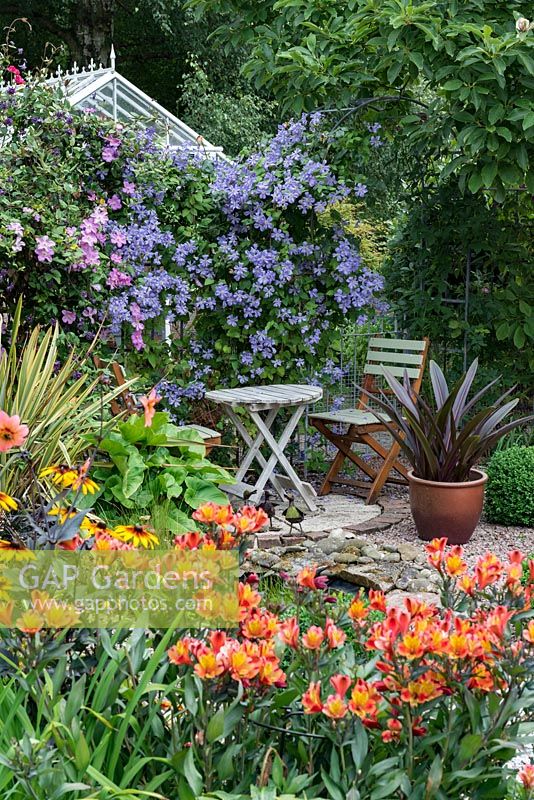 Table et chaises de bistrot en bois dans cour abritée, entourées de clématites fleuries 'Blue Angel' et Alstroemeria.