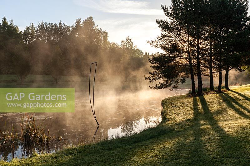 Brume s'élevant du lac avec sculpture en métal au lever du soleil. Plaz Metaxu Garden, Devon, Royaume-Uni.