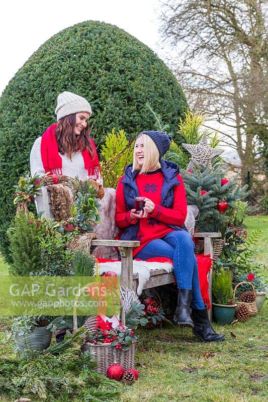 Femmes discutant par banc tout en buvant une boisson chaude, entourées de verdure et de décorations de Noël
