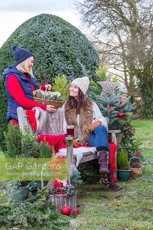 Femme donnant un panier avec des cadeaux à un ami assis sur un banc, entouré de verdure et de décorations de Noël
