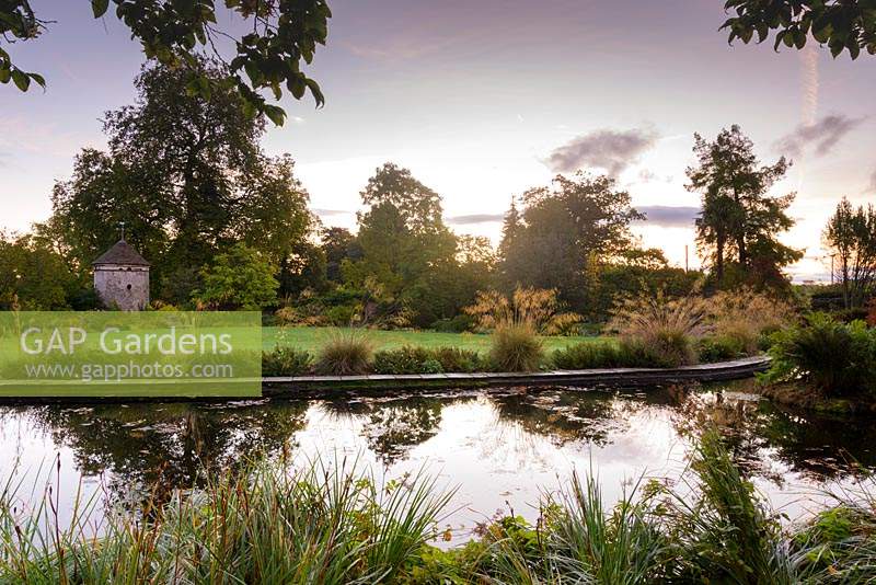 Un étang incurvé bordé de Stipa gigantea avec un pigeonnier au-delà, dans le jardin rond à Llanover Gardens, Monmouthshire, UK.