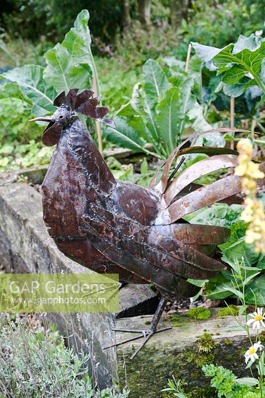 Sculpture de coq en métal assis sur la bordure végétale surélevée.