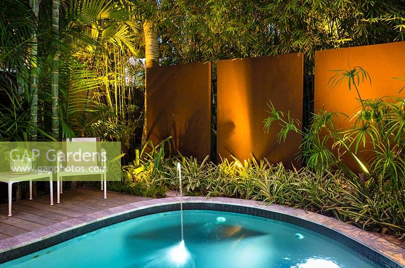 Jardin contemporain offrant intimité grâce aux murs et à la plantation tropicale. Conçu pour être utilisé la nuit avec éclairage, coin salon, terrasse en bois et piscine.