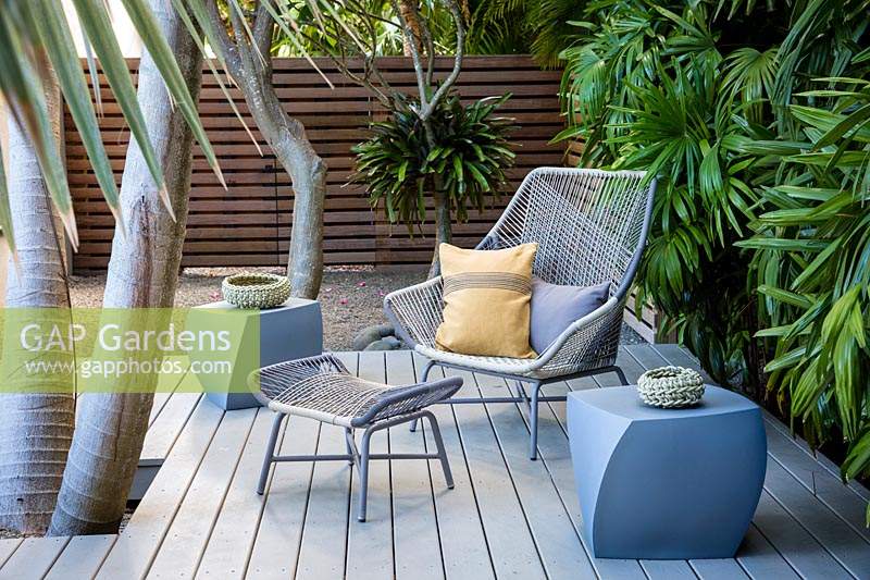 Espace détente sur terrasse en bois avec chaise, tabouret et petite table, terrasse en bois surélevée avec un trou taillé pour le tronc de palmier. Intimité assurée par les clôtures et le feuillage des palmiers