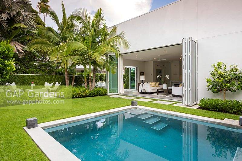 Vue sur la piscine à l'arrière de la maison avec ses portes pliantes ouvertes, au-delà des parterres de palmiers est un coin salon extérieur