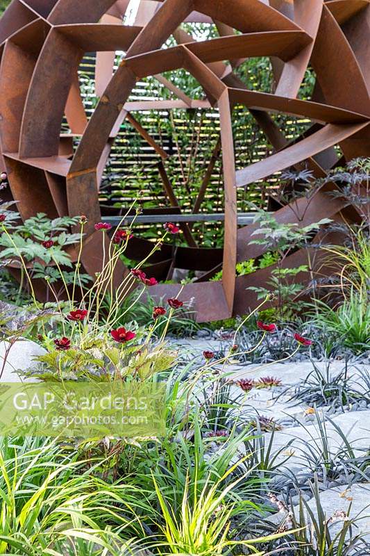 Sphère en acier corten de William Roobrouck près d'herbes ornementales en pavage dans 'Elements Mystique Garden', sponsorisé par Elements Garden Design