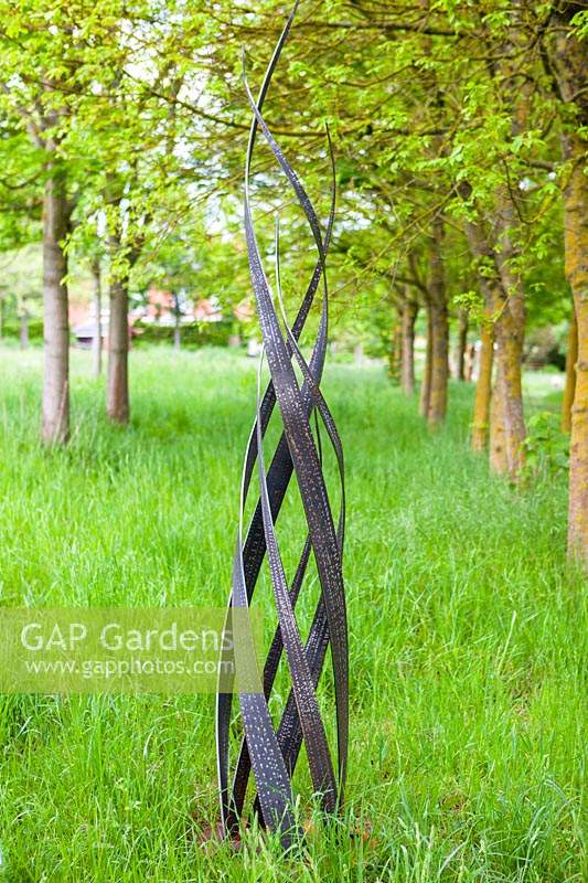 Flamme - sculpture en acier - dans l'herbe avec une allée d'arbres au-delà