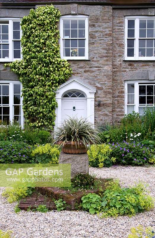 Zone gravillonnée avec socle central en pierre et plante en pot, avec vue sur la porte d'entrée entourée de parterres de fleurs vivaces. Bosvigo House, Cornwall, Royaume-Uni.
