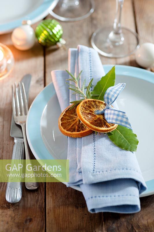 Créez vos propres ronds de serviettes en utilisant des matériaux naturels - des tranches d'orange séchées, des feuilles de laurier et du romarin attachées ensemble avec du ruban vichy bleu