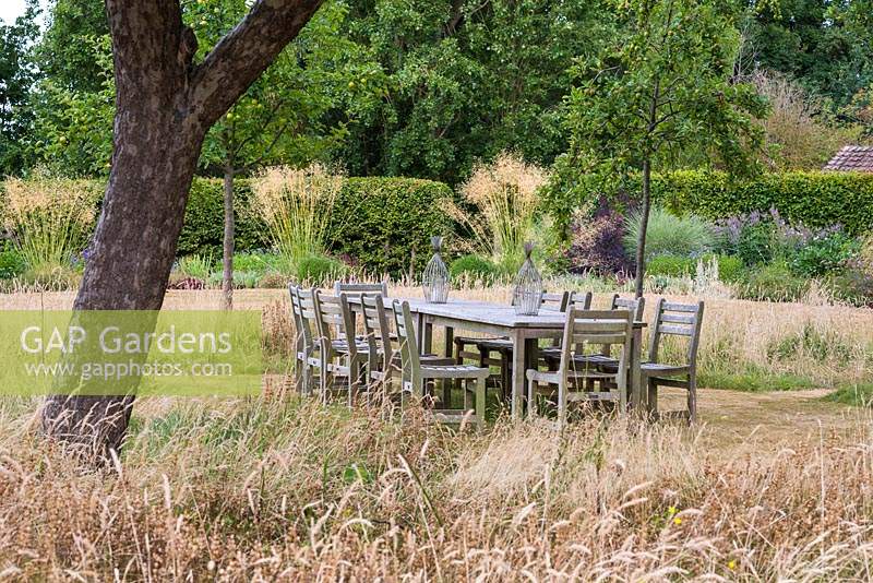 Table et chaises à l'ombre des pommiers, entourées d'herbe non coupée. Au-delà des parterres de fleurs parsemés de grosses touffes de Stipa gigantea.