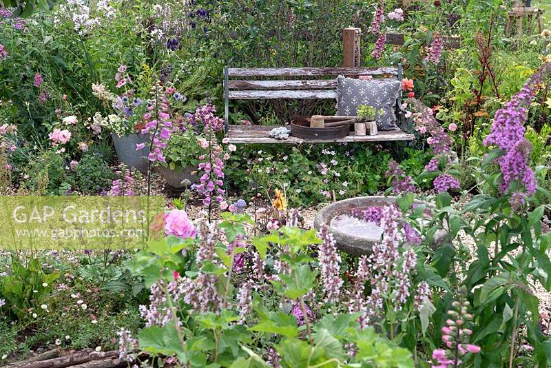 Outils de jardinage et coussin sur un banc de jardin rustique niché au milieu d'une plantation pérenne le long d'un chemin sinueux en gravier. RHS Hampton Court Festival 2019.