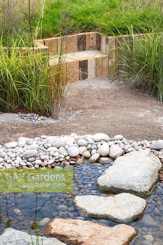 Un jardin de style naturaliste utilisant des matériaux naturels - un étang peu profond avec des cailloux et des pierres de gué, et une assise en bois installée dans un jardin en pente avec des herbes. RHS Hampton Court Palace Garden Festival 2019.