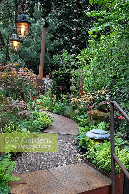 Vue du chemin en métal rouillé et déblais poreux de la carrière, bordé de geum 'Totally Tangerine' et d'herbes mixtes. Jardin: Carrière oubliée de Walker. Commanditaire: Walkers Nurseries. Chelsea Flower Show 2019.