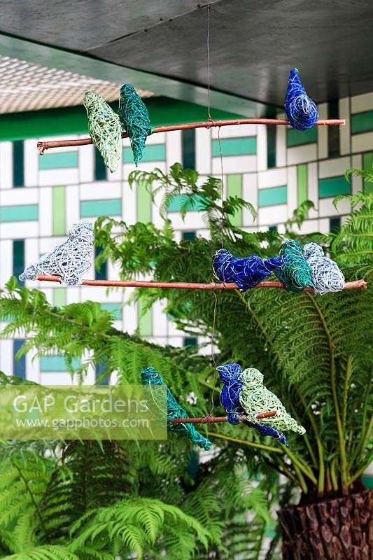 Le Jardin de bienfaisance Greenfingers. Mobile d'oiseau en fil vert et bleu avec Dicksonia Antarctica - Fougère arborescente et mur carrelé en céramique géométrique en vert et blanc derrière. Commanditaire: Greenfingers Charity