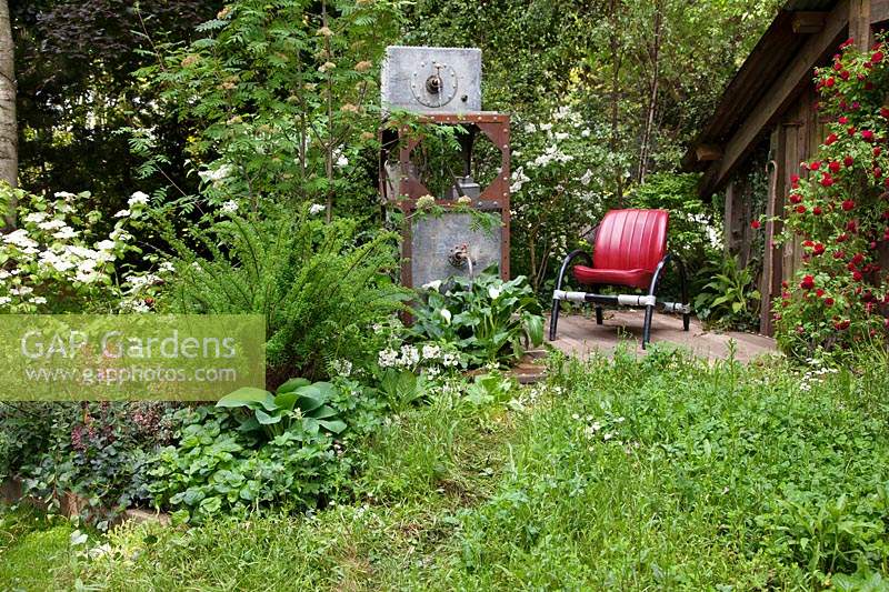 The High Maintenance Garden for Motor Neurone Disease Association, vue du jardin où de vieux pots en métal ont été recyclés en une pièce d'eau et où la plantation comprend des roses rouges errantes, Hosta, Viburnum et Primula - Sponsor: MND Association.