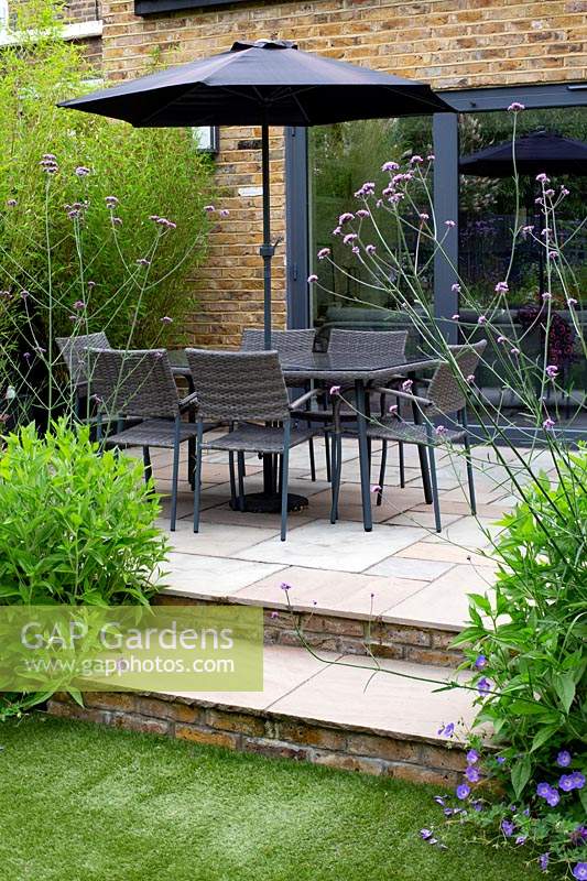 Jardin contemporain dans l'ouest de Londres - vue vers la maison et le patio en pierre avec table et parasol à travers les parterres de verveine bonariensis.
