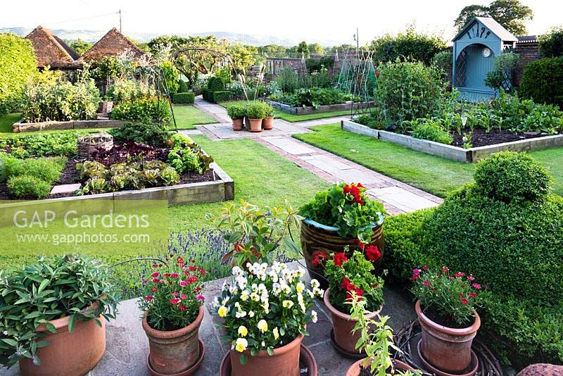 Vue du potager de la terrasse bordée de plantes en pot. Potager divisé en quartiers par des chemins pavés, chaque quartier a une pelouse avec de grandes bordures végétales surélevées bordées de traverses de chemin de fer, une tonnelle bleue le long de la clôture