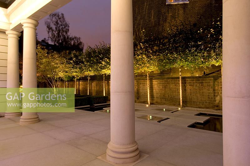 Vue à travers une terrasse pavée blanche avec des piliers sur une rangée d'arbres standard, éclairée par des projecteurs dans un jardin de ville contemporain.