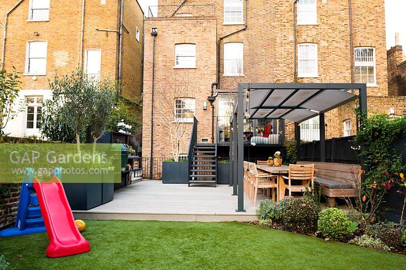 Jardin moderne avec aire de jeux, cuisine extérieure avec barbecue et coin repas avec de grands pots cubiques avec Olea europaea - Oliviers, Salvia officinalis et Betula pendula multi-tiges