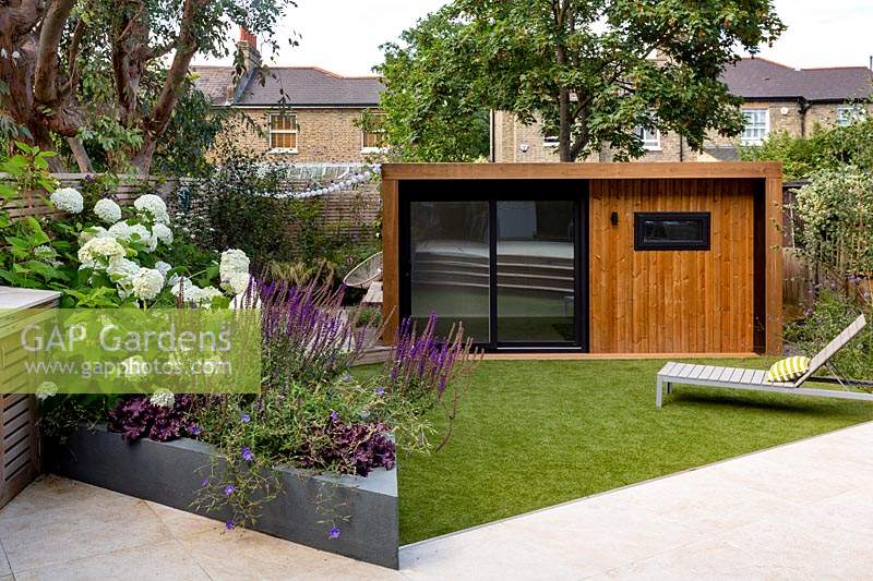 Jardin contemporain de Londres - vue sur le jardin de la maison vers la pelouse artificielle avec chaise longue, salle de jardin ou salle de gym et zone de terrasse en bois inférieure.