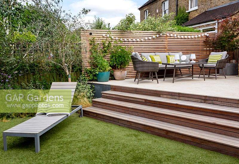 Terrasse de jardin contemporaine de Londres avec des marches en bois menant à un canapé de jardin moderne, des chaises et une table sur une terrasse avec une clôture en treillis de lattes de cèdre derrière. Au premier plan, une chaise longue moderne est assise sur une pelouse artificielle.