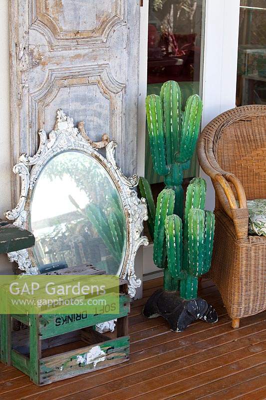 Une collection d'objets décoratifs sur une véranda, avec un cactus palster, une caisse de boisson gazeuse rétro, un miroir antique, une porte lambrissée et une chaise paon en rotin.