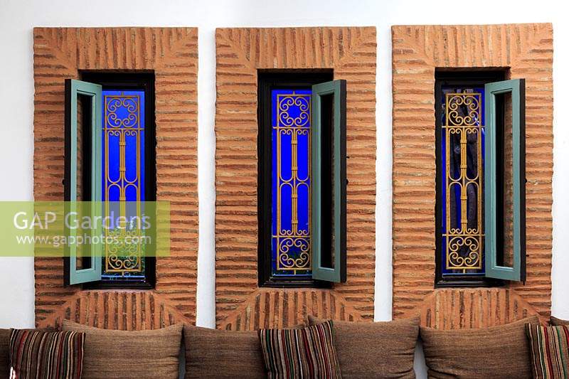 Grandes fenêtres étroites avec grille en métal ouvragé et mur bleu vif derrière, coussins sur chaise à l'avant. Le Jardin Majorelle, Jardin Majorelle, Marrakech