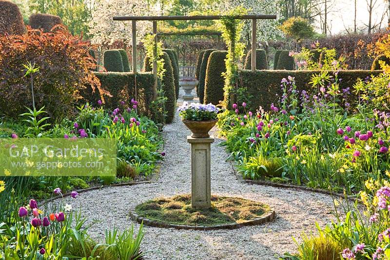 Jardin à la française avec urne centrale sur socle, parterres de bulbes à fleurs, colonnes topiaires au-delà