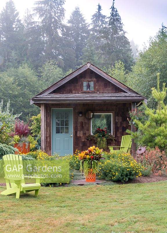Cabane rustique dans un jardin de campagne, décorée de jardins en pot orange et de chaises Adirondack chartreuse.