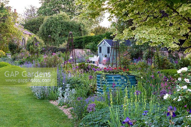 Un parterre de gravier est planté d'épis d'agneau argenté, d'alliums, d'iris et de centranthus, entourant un grand tambour métallique de dahlias rouges et de lierre. Au-delà, un pavillon d'été donne sur une terrasse et un parterre de fleurs herbacés ponctués d'obélisques roses.