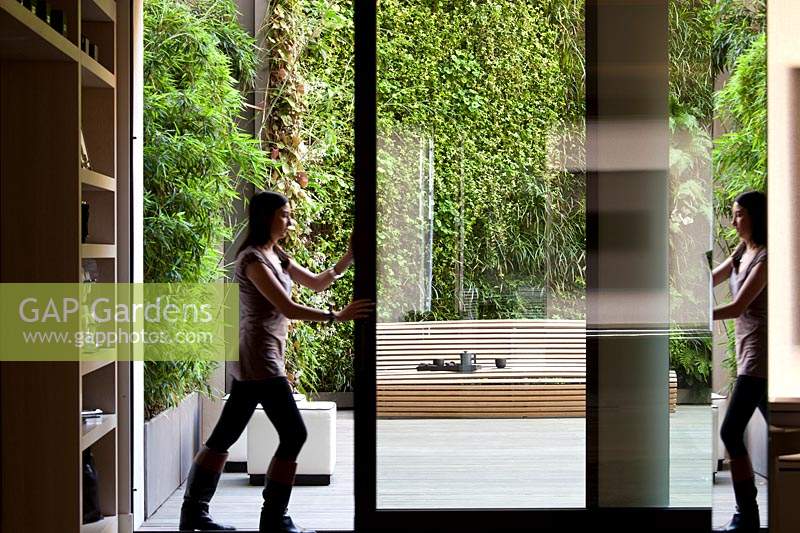 Femme poussant une porte moustiquaire ouverte pour révéler un jardin de cour moderne avec des murs végétalisés.