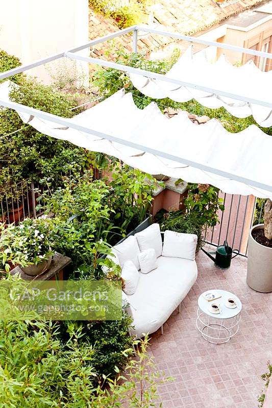 Petite terrasse carrelée vue de la maison, montrant un auvent ombragé sur un coin salon soutenu par des plantes en pot