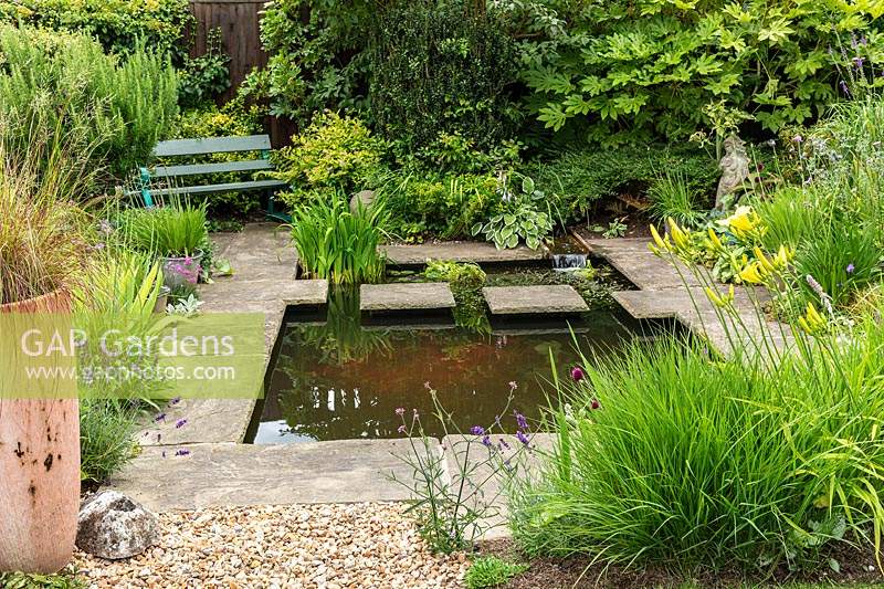 Un petit étang de jardin avec des tremplins dans un jardin de campagne de banlieue, entouré de pavés en pierre, d'un siège de jardin et d'une plantation mixte informelle de vivaces et d'arbustes