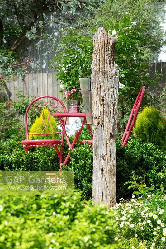 Une branche de bois rustique indépendante avec un pluviomètre en plastique monté devant une table et des chaises d'extérieur rouges.