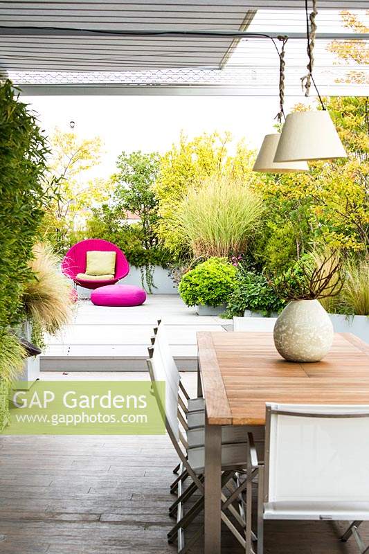 Salle à manger couverte sur terrasse blindée par des arbustes cultivés en pot et un espace de détente en arrière-plan avec une chaise et des coussins en osier rose vif