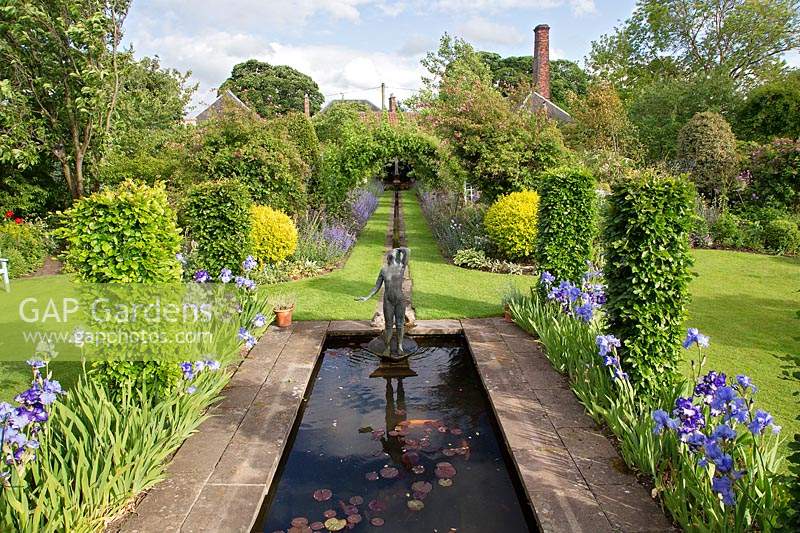 Étang rectangulaire avec parterres d'iris et fontaine sculpturale d'une jeune fille, dans une pelouse avec des parterres mixtes