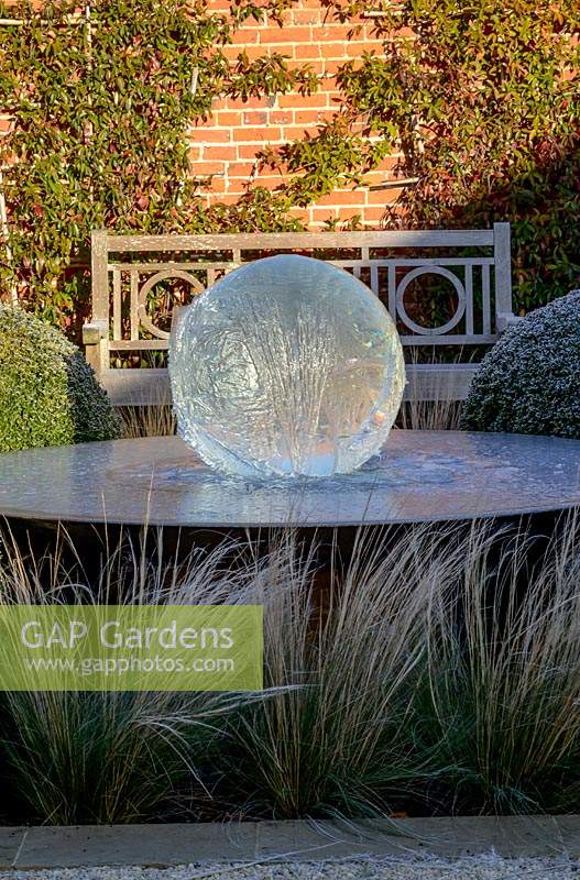 Pièce d'eau Aqualens conçue par Allison Armor avec de grosses boules coupées de Buxus sempervirens, de l'herbe Stipa tenuissima et de Traschelospermum jasminoides sur un mur de briques, un matin glacial.