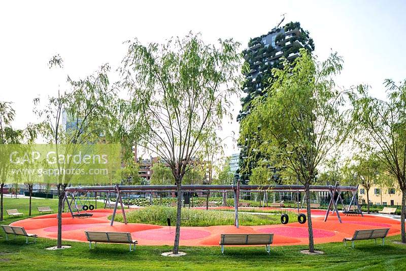 Vue sur le parc public avec de jeunes arbres et des bancs autour d'une aire de jeux, en arrière-plan le Bosco Verticale - forêt verticale - par l'architecte Stefano Boeri