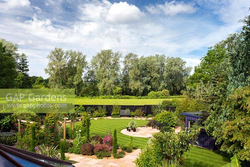 Vue d'ensemble du jardin avec patios circulaires, pelouse, parterres d'arbustes et clôture peinte en noir à la limite