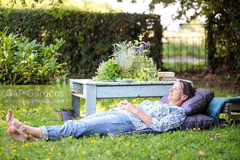 Femme reposante sur des coussins violets à côté d'une table en bois avec jardinière à herbes intégrée.