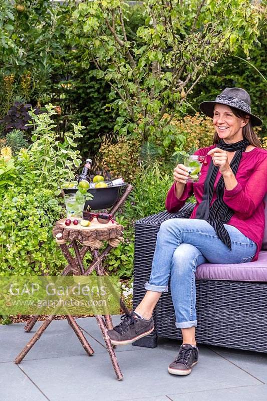 Femme se détendre et prendre un verre sur un patio en ardoise à côté d'un trio de structures de trépied - un planteur de menthe, une petite table de boissons et un refroidisseur de boissons