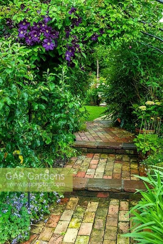 Chemin de brique à travers l'arche menant à la pelouse, avec Clematis viticella 'Etoile Violette' au-dessus - Journée des jardins ouverts, Double Street, Framlingham, Suffolk