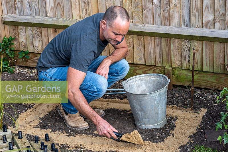 Après avoir nivelé le sol, l'homme asperge le sable doux du constructeur prêt à recevoir la couche inférieure de bois pour le parterre de fleurs surélevé.