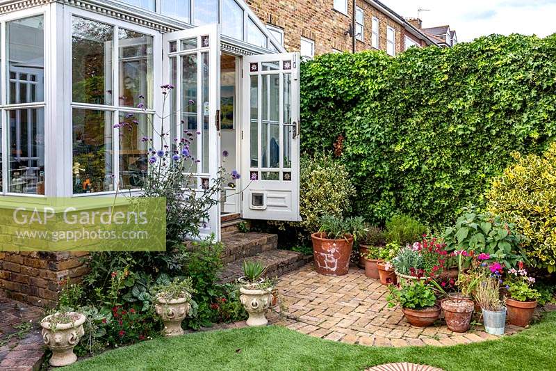 Petit jardin avec véranda attenante à la maison ouvrant sur un petit patio avec des plantes en pot