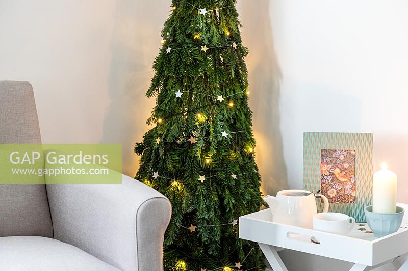 Sapin de Noël peu encombrant en pin - Abies et conifères - Feuillage de Juniperus attaché à un obélisque de saule et décoré de décorations et de lumières en étoile dans un salon moderne avec chaise et table d'appoint