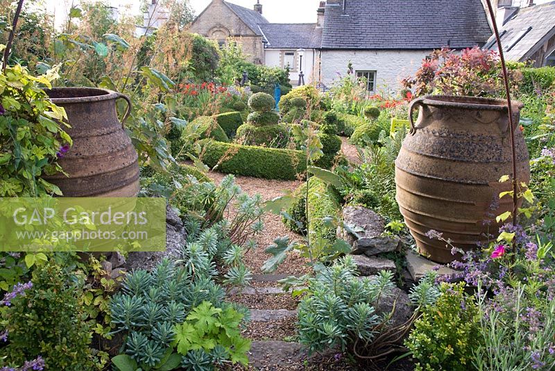 Vue d'ensemble du jardin de cottage avec topiaire, grandes urnes et parterres de fleurs informels.